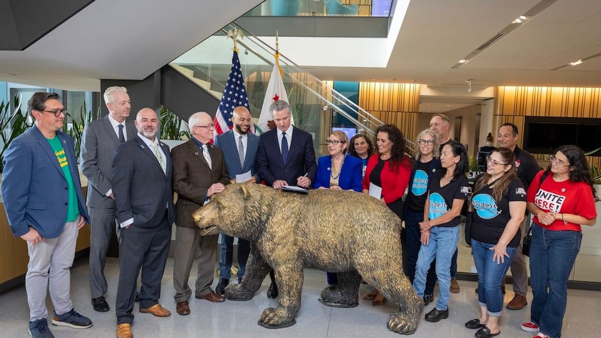 Governor Gavin Newsom signs worker protection bills beside a bronze bear statue, surrounded by legislators, labor leaders and workers
