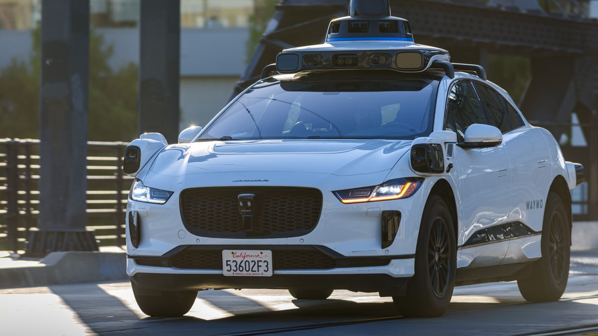 A white Waymo self-driving car with a California license plate and a ring of sensors on its roof