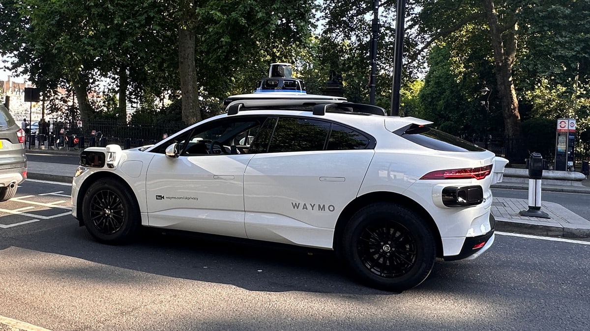 A white Waymo self-driving car with sensors on its roof on Northumberland Avenue in central London, beside a London bus stop sign