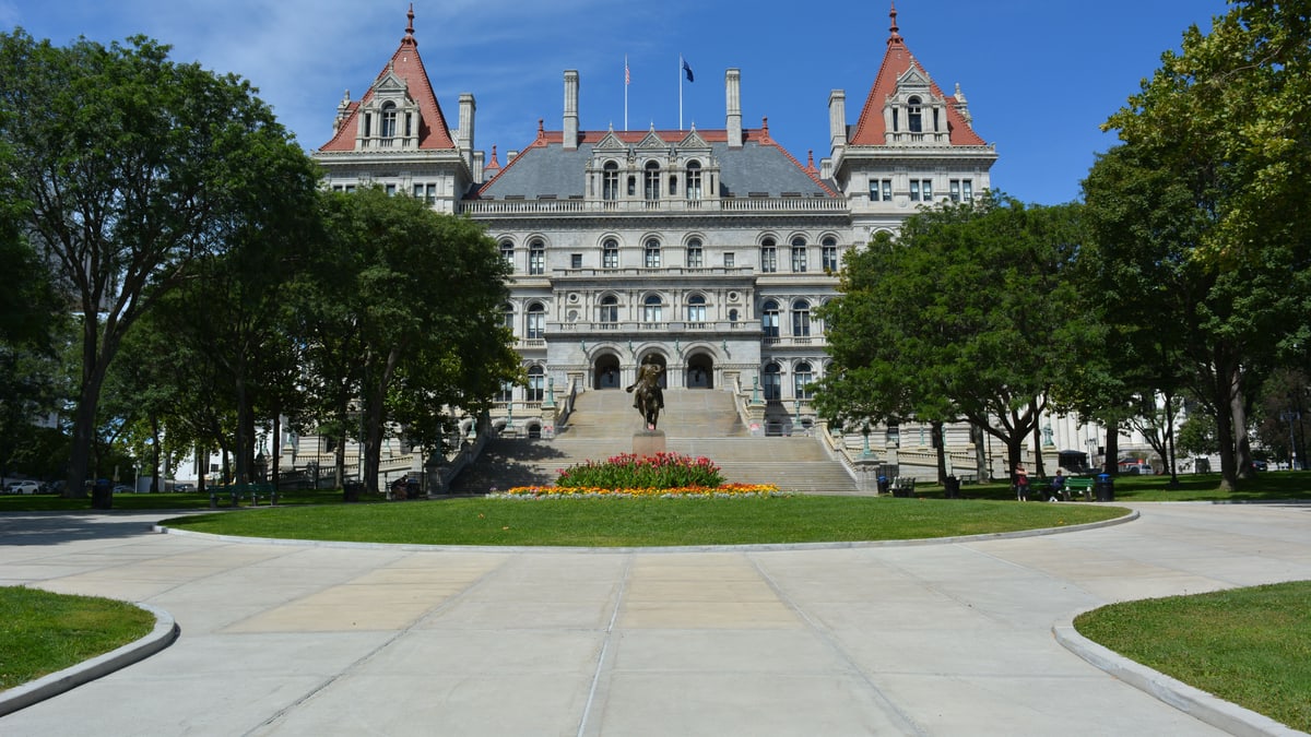 The New York State Capitol in Albany, a stone building with red-roofed towers, seen across a lawn and a flower bed on a sunny day