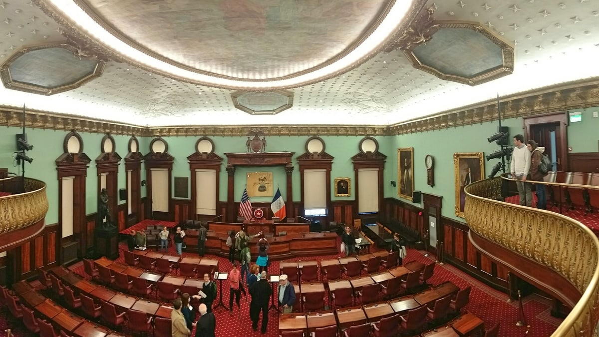 The New York City Council Chamber in City Hall, with rows of red chairs and desks facing the speaker's rostrum