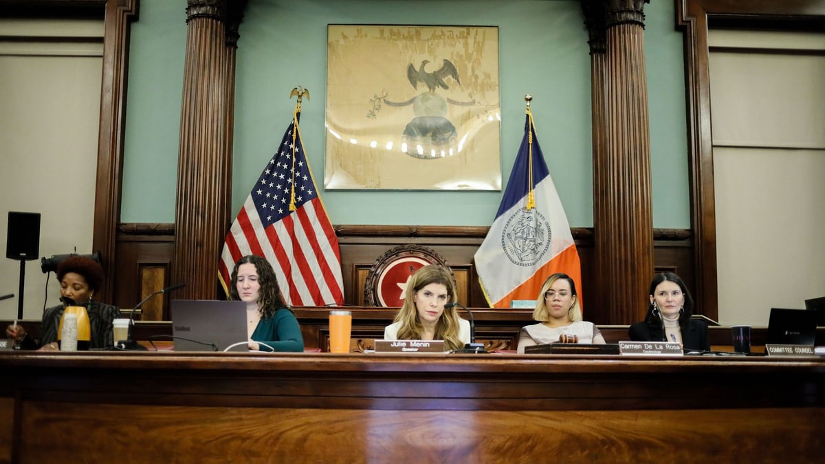 Speaker Julie Menin, Council Member Carmen De La Rosa and others at the dais of a New York City Council hearing room, between a U.S. flag and a New York City flag