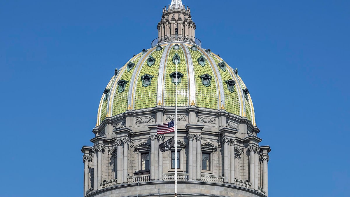 The green and gold dome of the Pennsylvania State Capitol in Harrisburg against a clear blue sky, with an American flag on a pole in front