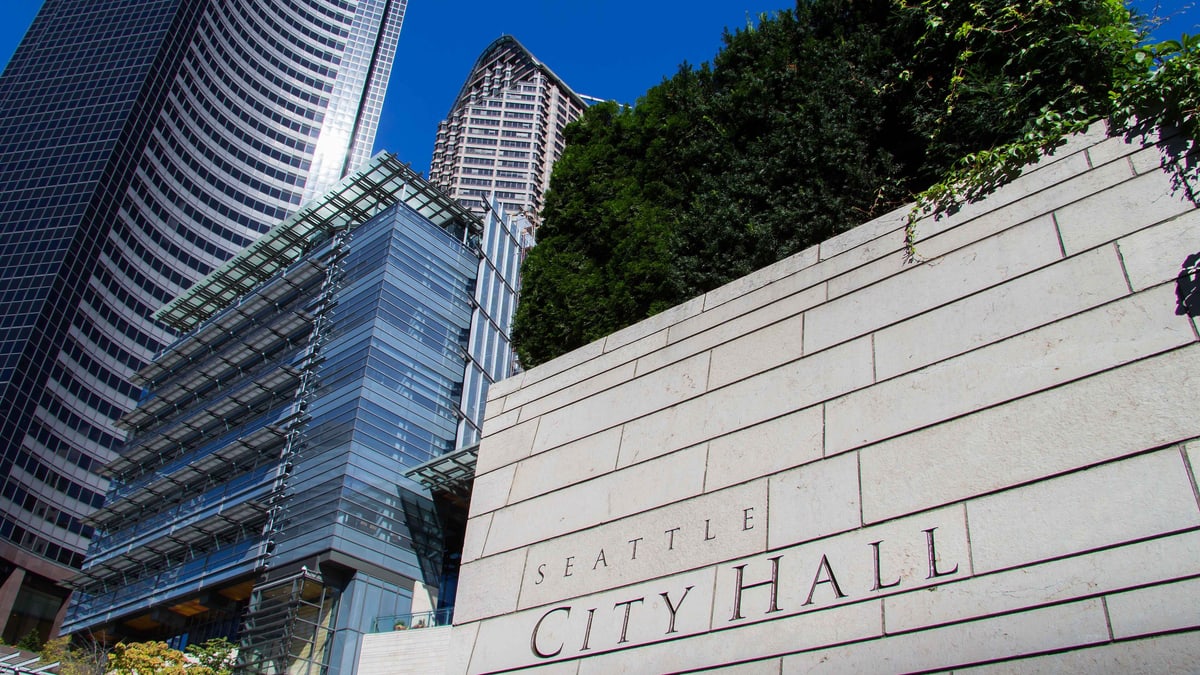 The words Seattle City Hall carved into a stone wall in front of the glass City Hall building and downtown towers