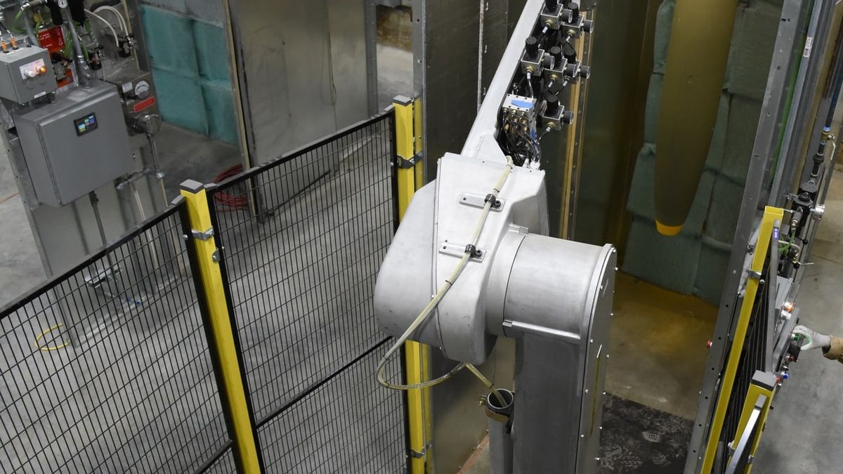 A large industrial robot arm inside a fenced cell at a U.S. Army ammunition plant, with a bomb body hanging in the paint booth behind it