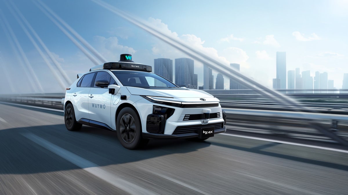 A Waymo image of a white Toyota bZ4X with Waymo's sensor dome on its roof, driving across a bridge with city towers behind
