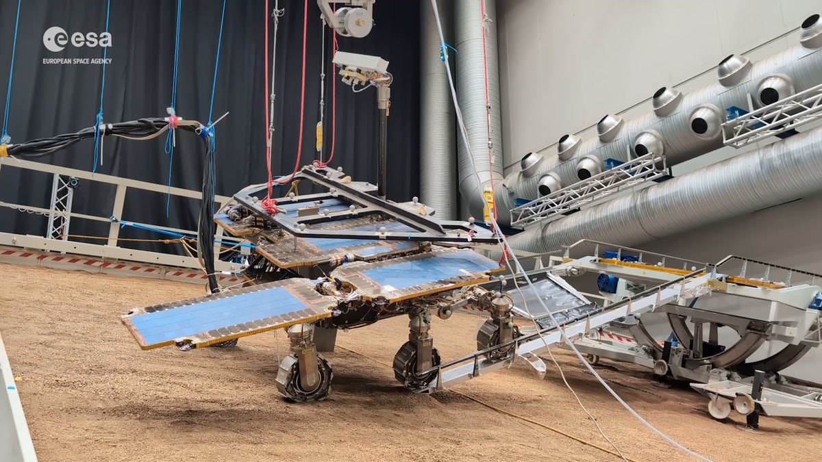 A full-size test model of the Rosalind Franklin Mars rover, held up by ropes from the ceiling, drives down a ramp from a mock landing platform onto reddish sand indoors