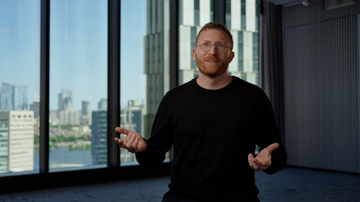 A man speaking in an office with a city view, beside a photo of four men talking around a laptop and the BIDMC sign on the hospital building
