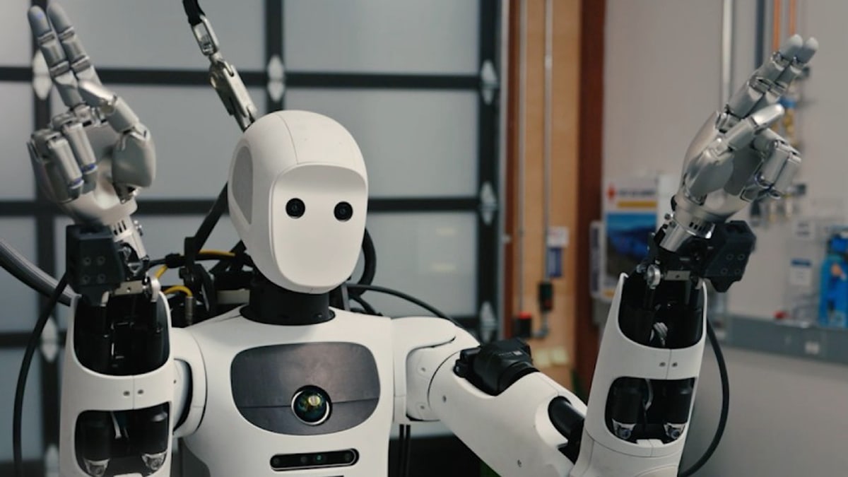 Apptronik's white Apollo humanoid robot raising both mechanical hands in a robotics lab, from Google DeepMind's cornhole video