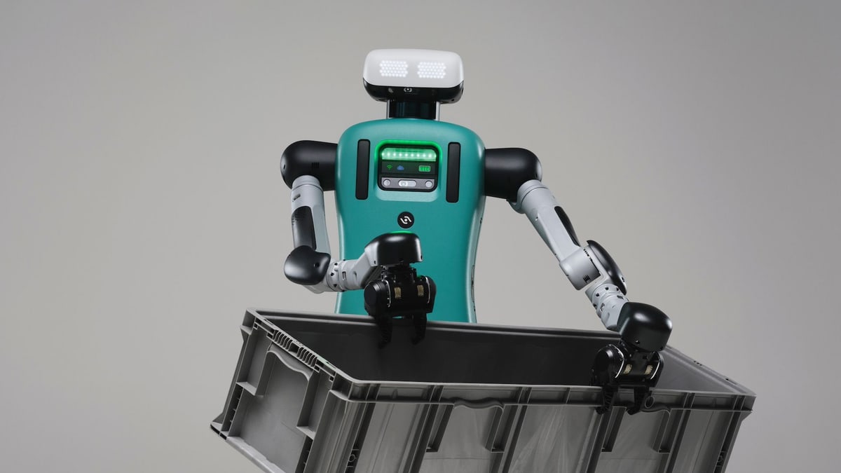 Agility Robotics' Digit humanoid robot, with a teal body and a white head, holding a gray plastic tote in both hands