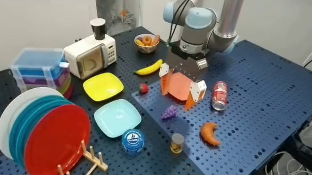 A robot arm with an orange gripper holds a pink plate above a blue table set with fruit, plates and a small toy microwave, in Kyutai's REKI test video
