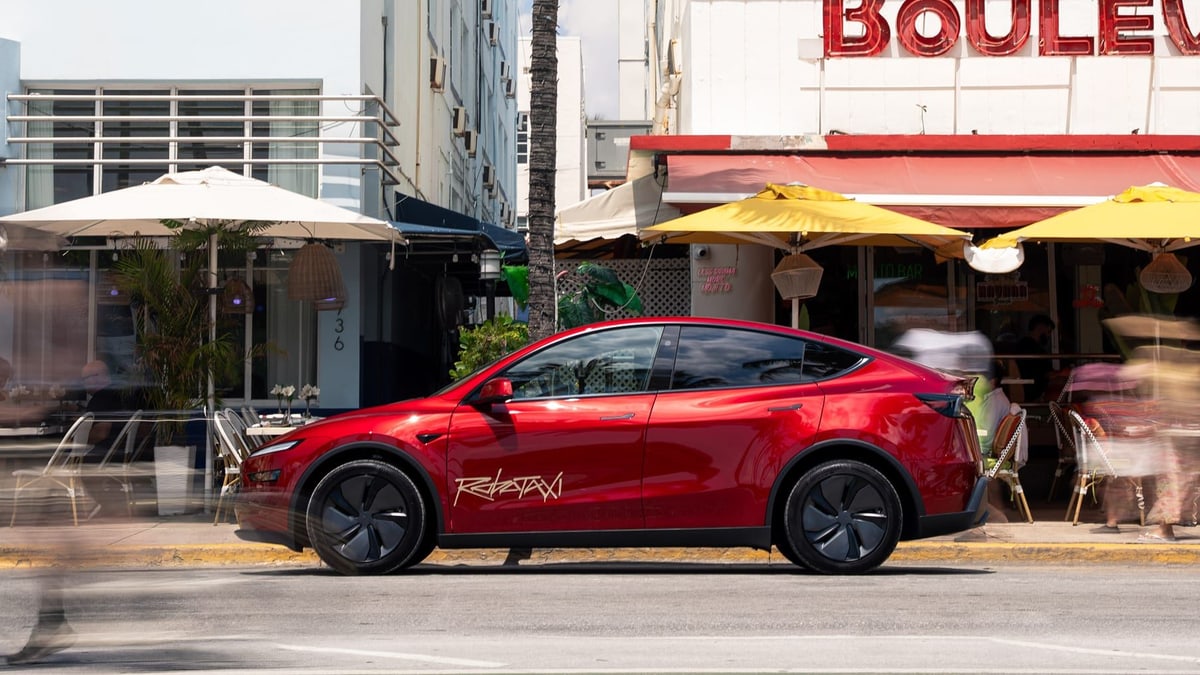 A red Tesla Model Y with Robotaxi lettering on its door, parked on a street lined with cafe umbrellas, from Tesla's Robotaxi page