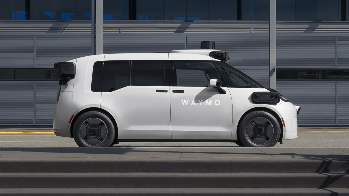 A white Waymo driverless minivan with roof sensors, seen from the side in front of a gray building