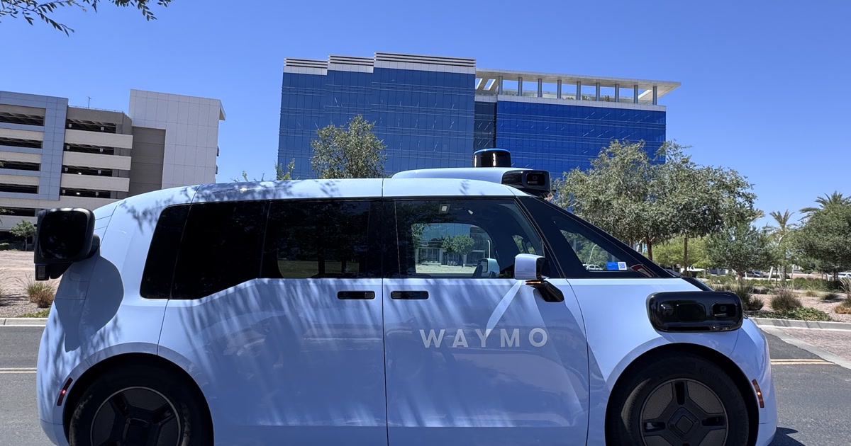 Light blue Waymo Ojai robotaxi with roof lidar parked beside an office building under clear sky